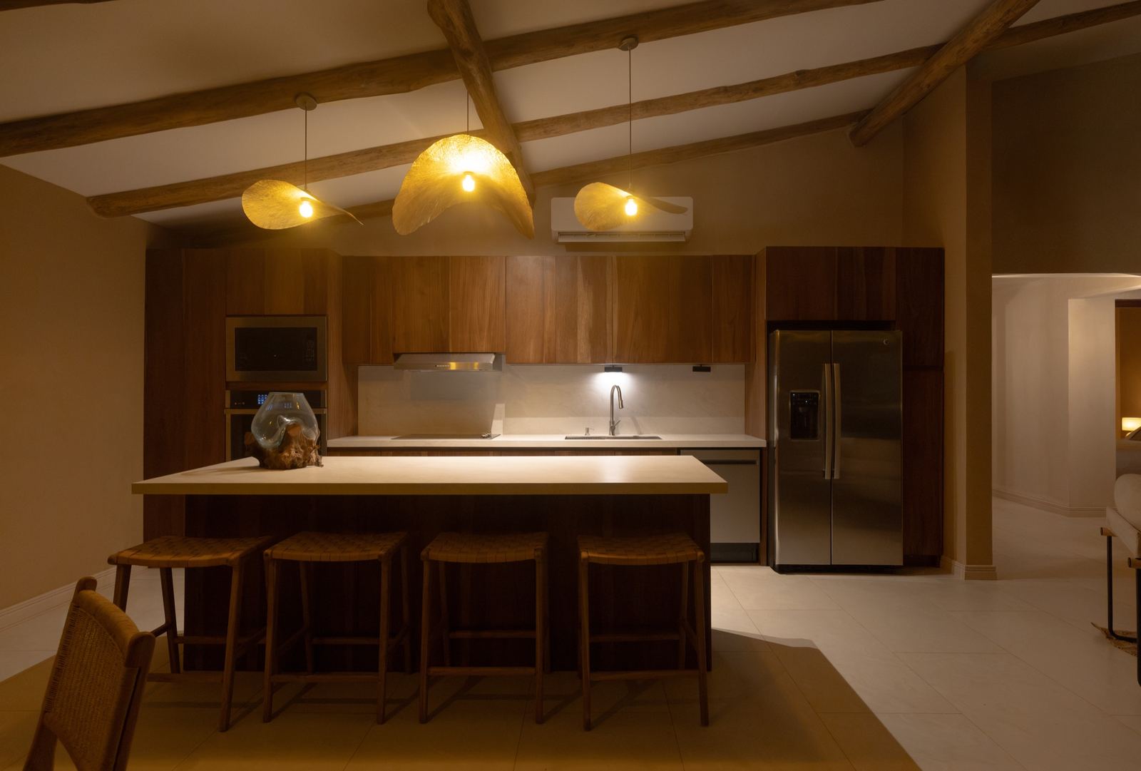 La cocina completa de noche: isla, cuatro bancos, refrigeradora y vigas de madera