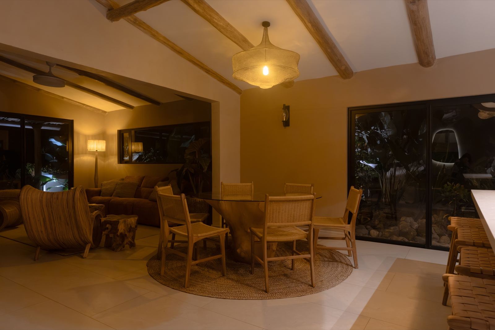 Comedor de Casa Bergamota 13: mesa sobre tronco, sillas de ratán y alfombra de fibra natural, con la sala al fondo